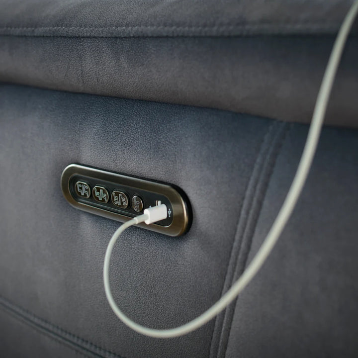 Close-up of a dark gray upholstered chair or sofa with an oval control panel mounted on the side, showing a white charging cable plugged into one of the ports.